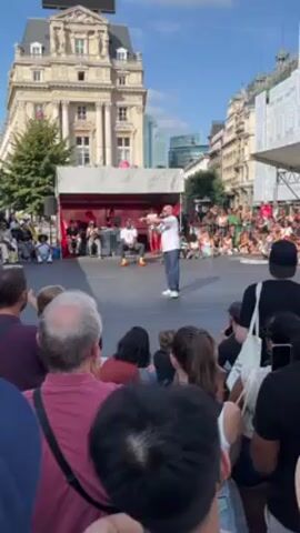 The video shows a man addressing a crowd from a stage. He's wearing a white shirt and dark pants. Behind him, under a canopy, people are seated. The audience is diverse, with people of different ages and ethnicities. The setting appears to be an urban square with buildings in the background, and the weather looks sunny.