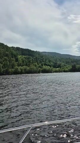 This is a first-person perspective video taken from the bow of a boat as it travels across a large body of dark water. On the horizon, there is a heavily wooded coastline with rolling hills. The sky is partly cloudy with patches of blue sky visible between large, white cumulus clouds. The water is choppy, with small waves reflecting sunlight. A silver railing is visible in the foreground, suggesting the camera is positioned on the deck of the boat. There are no people visible in the video.