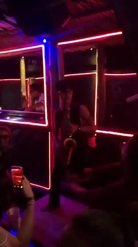 The video shows a bustling nightclub scene. A man is standing on a table, playing the saxophone for the crowd, while a DJ is visible in the background. People are dancing and enjoying themselves, some holding drinks or recording the scene with their phones. The club has a vibrant atmosphere, lit by neon lights and disco balls. A sign reading "Tequila Sunrise" hangs in the background.