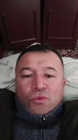 A man is lying down, wearing a gray turtleneck sweater and a dark jacket. He is positioned against a patterned pillow and what appears to be a bedspread. Behind him is a dark wooden cabinet with two doors. The man’s face is centered in the frame. He speaks. At one point he bares his teeth.