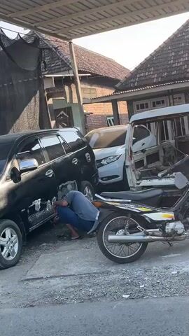 A person in blue pants and a dark shirt is crouched down, working on the side of a black car. The car is parked next to a white car and a motorcycle. The scene appears to be an outdoor repair shop or garage, with a metal roof overhead and buildings in the background.