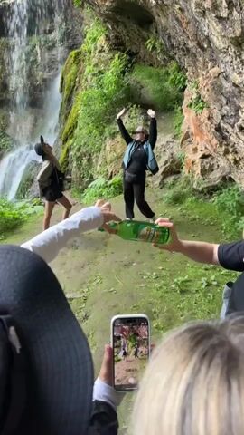 This video shows a group of people in a natural setting near a waterfall and a rocky overhang. In the foreground, two individuals are partially visible, one holding a smartphone and another holding a green glass bottle of beer. The smartphone is being used to record a video of the scene. In the midground, a woman is posed with her arms raised in the air, appearing to dance or strike a pose. She is wearing a black long-sleeved shirt, a teal vest, black pants, a baseball cap, and white sneakers. To her left, a person in a black t-shirt and shorts is also visible, holding a backpack and looking towards the waterfall. The ground is covered in moss and dirt, and lush green vegetation is visible on the rocks and in the background. The waterfall is cascading down a rocky cliff face. The overall atmosphere suggests a casual outing or a moment of fun captured on video.