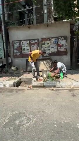 Two men are outside on a sunny day. One man, wearing a yellow shirt and gray pants, stands on a ledge near a small, public fountain while the other man, wearing a gray shirt, kneels near the fountain, filling a green plastic container with water. The fountain has a stream of water running from a pipe into a small basin. There is a building with posters on the wall behind them. There is a street in front of them.