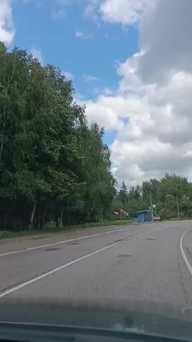 The video shows a view from a moving vehicle on a paved road. On the left side of the road, there is lush green foliage and trees. To the right of the road, there are several small houses with wooden roofs and blue siding, a blue bus stop shelter, a road sign indicating a curve to the right, and a lamppost. The sky is a bright blue with fluffy white clouds. The overall environment appears to be a rural or semi-rural setting.