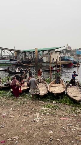 The video captures a bustling waterfront scene with several wooden boats docked along a muddy bank. Individuals are seen entering and exiting the boats, some using long poles to maneuver. One man is seated on a small wooden stool, facing the waterway. In the background, buildings and other watercraft can be observed. The overall atmosphere appears to be busy, likely a transportation hub or a commercial area along the water.