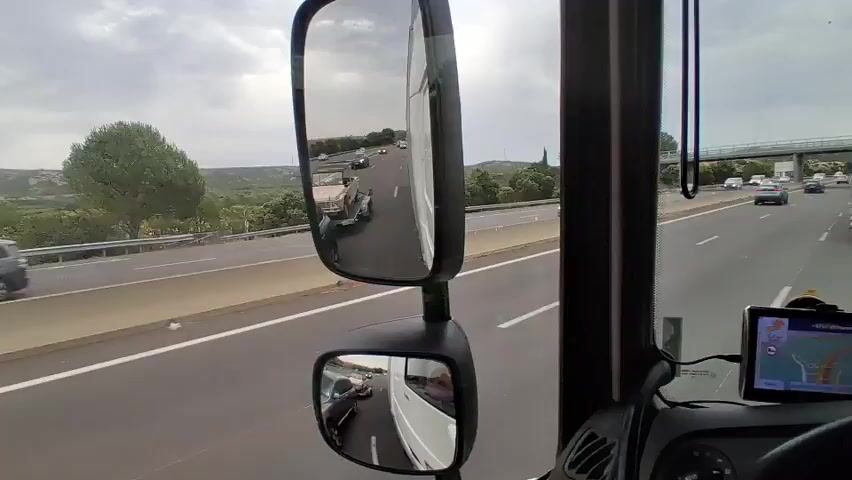 This media file shows a view from inside a vehicle, likely a truck or RV, driving on a highway. The highway appears to be multi-lane and moderately busy with other vehicles, including cars and trucks. The time of day seems to be late afternoon or early evening, with overcast skies suggesting potentially cloudy or drizzly weather. The surrounding landscape features rolling hills and vegetation, indicating a rural or semi-rural setting.

The primary focus of the video is the road ahead and the vehicles traveling on it. In the foreground, a small, light-colored vintage car is being transported on a trailer behind another vehicle. This vintage car has a canvas top and appears to be quite old. In the side mirrors, we can see reflections of other vehicles and the highway. The main side mirror shows a view of the highway with traffic moving in the same direction. The lower, smaller side mirror shows a closer view of the trailer and the vintage car.

There are a few instances of visible text. On the back of one of the trucks, there is a logo with a stylized face. On the trailer carrying the vintage car, there is a license plate which appears to be French. The GPS device mounted on the dashboard displays a map with a route indicated, showing "Lançon-Provence" as a destination or location.

The overall impression is a view of everyday travel on a highway, with a focus on the transportation of a unique vintage vehicle. There are no overt emotional cues, but the scene conveys a sense of steady movement and ordinary transit.