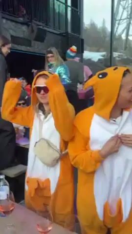 A group of people are celebrating while wearing orange and white animal onesies.  They are indoors, near windows, possibly at a ski lodge.  Some of them are holding wine glasses.  They are laughing and dancing.  Other people are in the background, and some are not wearing the onesies.  There is snow visible outside.