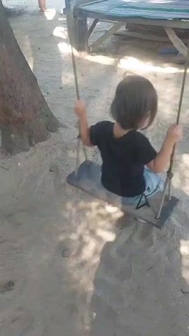 The video shows a child on a swing. The child is wearing a black t-shirt and blue shorts. The swing is made of wood and rope. The child is holding onto the ropes of the swing. The swing is in motion, and the child is swinging back and forth. The background of the video is a sandy beach. There is a tree to the left of the swing, and a wooden structure to the right. Sunlight is casting shadows on the sand.
