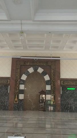 The video pans across the interior of a mosque in Tilamuta, Indonesia. The scene is well-lit, suggesting it is daytime. The camera begins by focusing on the ornate mihrab, a semicircular niche in the wall that indicates the direction of Mecca. The mihrab is decorated with intricate Arabic calligraphy on a dark brown background, framed by a marble border. Around the mihrab, there are alternating white and black marble segments.

To the left of the mihrab, a white, coffered ceiling with a simple light fixture is visible. To the right of the mihrab, the floor is tiled with long, grey rectangular tiles, and a dark marble wall is present. A digital display with green text, reading "ADZAN SHUBUH", is mounted on this wall, indicating it is time for the dawn prayer. A small lectern is also visible within the mihrab area, likely for reciting prayers or reading from the Quran. The overall impression is one of a clean, well-maintained, and spiritual space. No people are visible in the video.