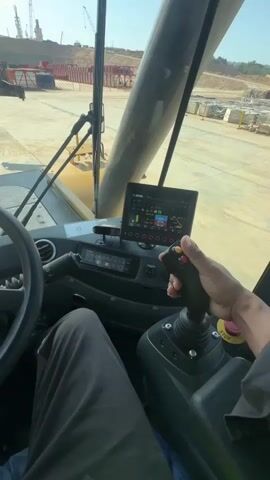The video shows the interior of a crane cabin. A person's hand is visible operating a joystick control. The joystick appears to be a multi-functional control with buttons and a trigger. A digital display screen is mounted on the dashboard, showing various operational data and graphics, likely related to the crane's status and functions. A steering wheel is also visible to the left of the cabin. Through the front window of the cabin, an outdoor construction site can be seen with heavy machinery and materials. The overall environment suggests ongoing construction work. The video is shot from a first-person perspective, as if the viewer is inside the crane cabin.