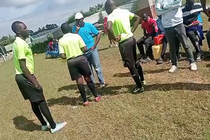 The video shows a group of people, likely referees, coaches, and team staff, gathered on a grassy field near a building. Three referees are wearing matching neon yellow shirts and black shorts, one person is wearing a light blue shirt and jeans with a white baseball cap, and other people are wearing various athletic apparel. They appear to be in a discussion. Some of the people are standing, while others are sitting on benches nearby. The environment suggests a sports field, possibly a soccer field. The sky is partially cloudy.