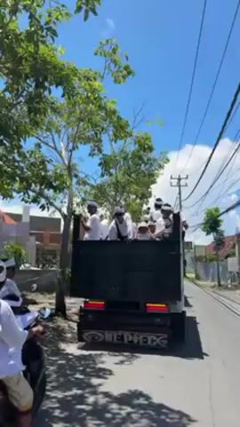 A dump truck with approximately eight people is seen moving down a road on a sunny day. The people are wearing white clothes and hats. The dump truck is black and has writing on the back. The road is paved and there are trees and buildings on either side of the road. A person on a scooter is visible in the bottom left of the frame.