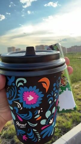 This is a close-up, eye-level shot of a person's hand holding a black paper coffee cup with a colorful floral design. A small, green candy bar with text in Russian is held alongside the cup. The cup and candy are the primary subjects, positioned in the foreground.

The background reveals an outdoor scene with a grassy field and a row of apartment buildings in the distance. The sky is blue with some clouds, and a faint rainbow or atmospheric halo is visible. The lighting suggests it is late afternoon or early evening, with the sun low in the sky.

The visible text on the cup is "Ярче!" which translates to "Brighter!". The text on the candy bar includes "babyfox", "DUBBLE CHOCOLATE", and several lines of Russian text describing the product as "Milk chocolate with crispy pistachio filling".

The overall impression is one of a casual, relaxed moment outdoors, possibly during a walk. The bright colors on the cup and the sunlight contribute to a positive, cheerful mood. The setting appears to be a suburban or semi-urban environment in Фролы, Russia, indicated by the apartment buildings and the Russian text. No people are clearly visible in the frame, though a distant figure can be seen walking in the field.