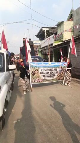 A group of people are participating in a parade. A woman in traditional clothing, holding a large flag, walks alongside a pickup truck. The truck has a banner on it that reads "ROMBONGAN KERAJAAN REMBANG RT 006/02 Desa Tanahbaya". Several other people are on the truck, some holding flags and others appearing to be part of a performance or display. Buildings line the street, and decorative banners are visible on some of them. A young boy in a black t-shirt and pants is walking in the foreground, facing away from the camera. The overall scene suggests a cultural or celebratory event.