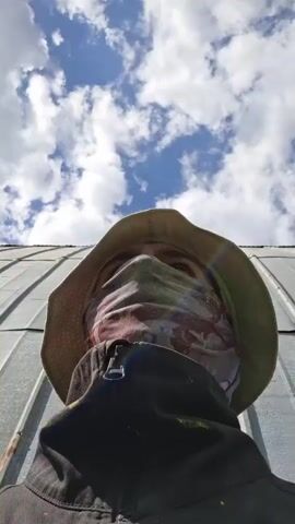 The video shows a person from a low angle, wearing a khaki bucket hat, a patterned face covering, and a black jacket. Above the person is a corrugated metal roof, and beyond that, a blue sky with scattered white clouds. Towards the left side of the frame, a pair of work gloves moves in the air. The sound of train cars can be heard in the background.