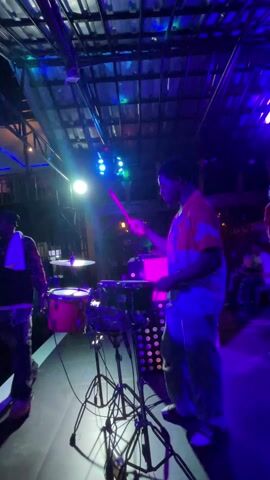 The video captures a live music performance indoors, likely at a club or event venue in Obosi, Nigeria. The central subject is a young man, possibly a teenager or in his early twenties, skillfully playing a drum kit. He is dressed casually in a patterned shirt and light-colored pants.

The drum kit includes a snare drum, cymbals, and a bass drum, along with an electronic percussion pad. The drummer is actively using drumsticks to hit the various components of the kit. His facial expression indicates focus and engagement in the music.

The setting is characterized by dynamic and colorful stage lighting, which frequently changes throughout the video. The lights shift from blue to green, then to purple, and at times incorporate yellow or orange hues, creating a vibrant and energetic atmosphere. These changing lights illuminate the drummer and parts of the stage, highlighting the performance.

In the background, hints of other band members or audience members can be seen, partially obscured by the lighting and stage setup. The overall scene suggests a lively and entertaining musical event taking place at night. There is no discernible text visible in the video.