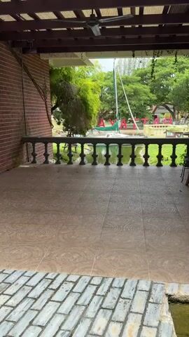 A person's perspective is looking out from a covered patio. The patio floor is tiled with a circular pattern. In front of the patio is a white railing with decorative balusters. Beyond the railing is a body of water, possibly a canal or pond, with a white sailboat docked in it. There are trees and greenery surrounding the water, and in the background, large red letters spelling "AYATHAI" are visible. The sky is overcast.