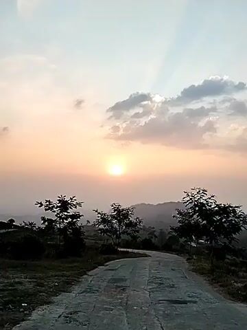 A paved road curves through a grassy, hilly landscape with scattered trees. The sun is setting, casting a warm glow across the sky, which is dotted with clouds. The scene appears to be a rural or park-like setting during dusk.