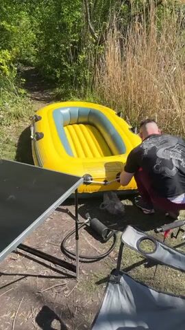 The video shows a person preparing for a fishing trip near a lake. Initially, a man is inflating a yellow and grey inflatable raft using a pump. The raft is on the grass next to a folding table and chair. The camera then pans to show the raft floating in the lake, with fishing rods placed beside it. Following this, a collapsible wagon is seen parked near a tree, filled with fishing equipment. A large bag and other fishing gear are laid out on the ground near the wagon. Finally, the camera shows multiple fishing rods set up on tripods, with their lines cast into the water. The environment is sunny, with grass, trees, and reeds bordering the lake.