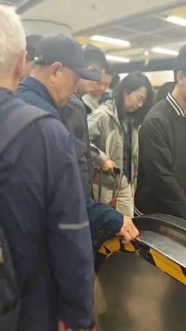 The video shows a busy indoor public space, likely a subway station or transport hub, in Fengtai District, China. Numerous individuals are seen boarding and riding an escalator.

In the initial frames, the camera focuses on a group of people at the bottom of the escalator. An individual with gray hair and a blue jacket with a backpack is in the foreground, facing away from the camera. Next to them, a person wearing a dark blue baseball cap is visible. Further ahead, a person in a light grey jacket and glasses is seen holding onto the escalator handrail.

As the video progresses, the camera pans upwards, revealing a longer section of the escalator. Many people, including adults and possibly children, are ascending. They are dressed in various casual attire, including jackets and backpacks. One person's black jacket clearly displays the text "CONVERSE ALL STAR" on the back. The escalator has metal and glass railings with yellow and black safety markings at the entry point. The lighting is bright and artificial, typical of an indoor public facility. The overall atmosphere appears orderly, with people moving steadily up the escalator.