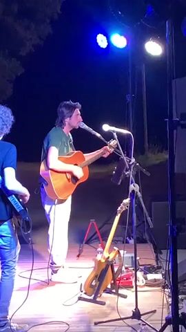 A medium shot captures a male musician playing an acoustic guitar and singing into a microphone on a dimly lit stage at night. He wears a green t-shirt and white pants. Another musician, visible from the side and back, plays a guitar. The stage is illuminated by blue spotlights and other stage lighting. Various musical equipment, including microphones, guitar stands, pedals, and speakers, are visible on the stage. The background is dark, suggesting an outdoor evening performance.