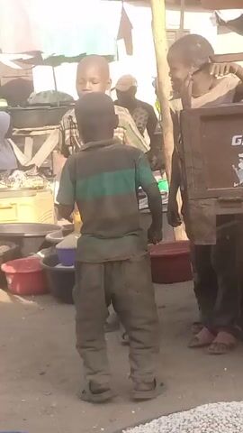 The video shows a group of children standing in what appears to be a market or street vending area. There are at least three children, one wearing a green and grey striped shirt and dusty pants, another in a plaid shirt, and a third with glasses leaning against a wooden structure that may be part of a vending cart. The first child is holding a black bowl or container. The environment is cluttered with various items, including red and black tubs, sacks, and other containers. There are stalls and overhead coverings, suggesting an outdoor market setting. Other people are visible in the background, including a woman in a hijab. The overall impression is one of a bustling, possibly impoverished, area.