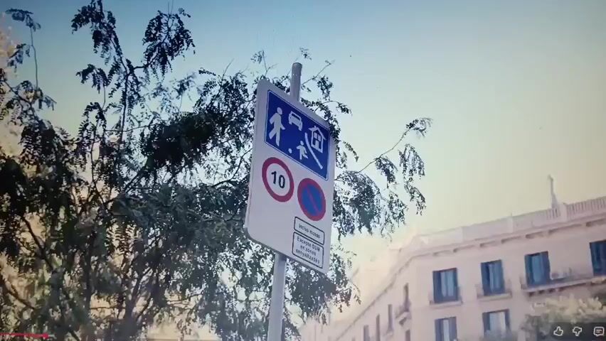 The video begins with a close-up of a street sign. The sign has multiple symbols on it, including a pedestrian crossing, a house, a car with a line through it, and a circle with a number 10 inside. Below these symbols, there is text in Spanish. The camera then pans out to reveal a street scene. There are trees lining the street, with buildings in the background. Several people are walking on the sidewalk and crossing the street. A white car is parked on the side of the road. The scene appears to be in a city, possibly in Europe, given the style of the buildings and the Spanish text on the sign. The lighting suggests it is daytime, with the sun casting some shadows. The overall atmosphere seems to be one of urban activity.