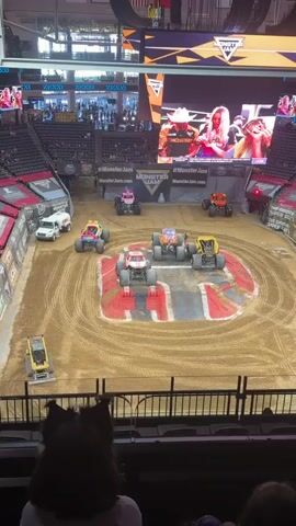 The video shows a monster truck rally. Several monster trucks are in a dirt arena. One truck is red and white. Another truck is purple and orange. Another is yellow. A pink truck with a unicorn design is visible. A yellow and blue truck is also present. A white truck with a tank is parked on the side. A yellow tractor is on the dirt. People are sitting in the stands, watching the event. A large screen at the top of the arena displays images of people. The event appears to be Monster Jam.