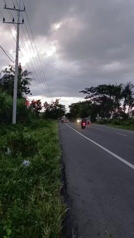 The video shows a two-lane paved road with vehicles traveling in both directions. A white line divides the lanes. The right side of the road has a grassy verge with dense vegetation and trees. A utility pole with overhead wires stands along the side of the road. The sky is overcast with dark clouds, and the sun is partially visible. The lighting conditions indicate it is late afternoon or early evening. The audio includes engine noise from the passing vehicles.