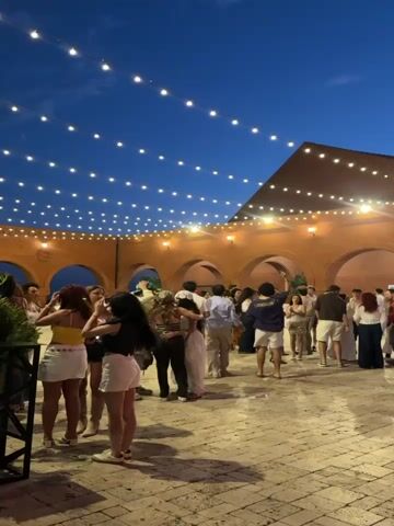 The video shows a gathering of people in an outdoor courtyard at dusk. String lights are hung across the open space, illuminating a group of individuals socializing. The architecture suggests a Spanish or Mediterranean influence, with arched walkways and earthy-toned walls. The people are dressed casually, and some appear to be dancing or mingling. The sky is a deep blue, indicating the transition from day to night. The ground is paved with stones.