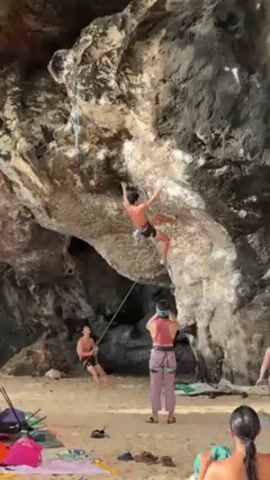 A man is rock climbing on a rocky overhang on a beach. He is secured by a rope, with another man sitting on the sand to hold the other end of the rope. A woman wearing a harness stands near the seated man. Two additional people stand in the background, near a pile of various items on a blanket. The rocky overhang is light gray in color, with dark gray crevices, with small white hand holds placed on the rock. The sand is light tan. The climber is wearing black shorts. The seated man is also shirtless. The woman wears a pink halter top and lavender pants.