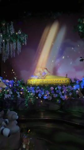 A nighttime scene unfolds with a dramatic lighting effect. A golden platform, adorned with purple flowers and shimmering lights, serves as the centerpiece. A figure, appearing to be a woman in a long yellow gown, lies on the platform, bathed in a golden spotlight that beams down from above. Next to her, a man in a white and blue outfit kneels, seemingly embracing or kissing her. Surrounding the platform is a lush, dark environment with green foliage and more glowing blue and purple flowers. Wisps of smoke or mist add to the ethereal atmosphere, with small lights twinkling in the background like stars. In the foreground, the back of a person's head is visible, wearing pink and white striped ears, suggesting they are an audience member at an event. The video pans slightly, offering glimpses of the surrounding dark, possibly wooded, environment.