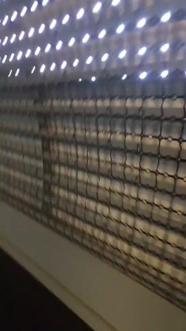 The video begins with a close-up of a patterned surface composed of small lights against a dark, grid-like background. The camera then pans to reveal a curved, tunnel-like structure with the same illuminated patterned surface forming the ceiling or upper walls. In the center of the frame, a reflective, disc-shaped sculpture on a stand is visible. The sculpture has a golden center and reflects the surrounding lights and the curved architecture. Light streaks are cast on the dark floor from the sculpture. The camera continues to pan, showing more of the illuminated patterned walls and the reflective sculpture. The environment appears to be indoors, possibly a public space or an art installation, with a dark, polished floor.
