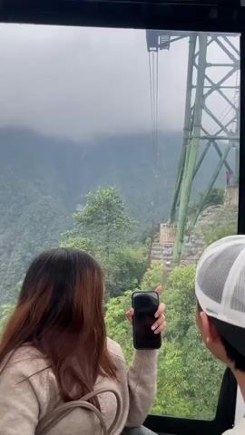 A person with long brown hair and a beige sweater is in the foreground, facing away from the camera and holding a black smartphone. To the right, a person wearing a white and black baseball cap is also visible from behind. They are inside a cable car, looking out at a misty mountain landscape. In the distance, a green cable car tower supports cables that stretch across the frame. Several cable cars, including one red and white car, are visible along the cables. The mountain slopes are covered in lush green vegetation, and the sky is overcast and cloudy. The person with brown hair raises their hands and waves towards the cable cars in the distance.