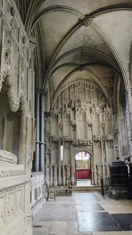 The video shows the interior of a large stone building, likely a cathedral or church, with ornate architectural details. In the foreground, a large stone monument with intricate carvings and a sculpted figure lies on its side. The camera pans to reveal massive stone columns and arches supporting a vaulted ceiling. Sunlight streams in through large stained-glass windows in the background, illuminating the space. Two people are visible in the distance, one standing and looking at the monument, and another seated near the windows. Informational signs are placed on the floor. The overall impression is one of historical grandeur and architectural magnificence.