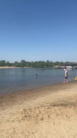 A wide shot shows a beach and a body of water under a clear blue sky. On the sandy beach in the foreground, a person wearing a white cap and blue shorts stands facing the water, and another person in yellow is sitting down. In the water, a person swims, and further out, two people are on paddleboards. Lush green trees line the opposite bank of the water, and a bridge is visible in the distance. The overall scene depicts a sunny day at a recreational water area.