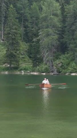 A video clip shows a couple rowing a wooden boat on a calm, green lake. The boat is in the center of the frame, and the couple is facing away from the camera, rowing in unison. They are wearing light-colored clothing. The lake is surrounded by a dense forest of tall, green pine trees, with a steep, wooded hillside forming the backdrop. The water is smooth, with subtle ripples reflecting the light. The overall scene evokes a sense of tranquility and natural beauty. The weather appears overcast, with soft, diffused light, suggesting it might be a cloudy day or early morning/late evening. There is no visible text in the media file. The scene is set in a natural landscape that is consistent with the Auronzo di Cadore region of Italy, known for its picturesque lakes and mountain scenery. The activity is a leisurely row on the lake, suggesting a peaceful outing. The emotions conveyed are those of calmness and perhaps romance or shared enjoyment of nature.