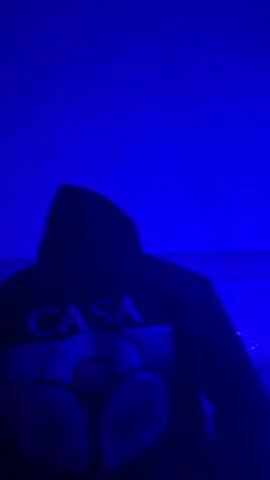 A person in a black hooded sweatshirt, with the word "CASA" printed on the back, stands facing away from the camera. The background lighting changes rapidly from green to blue to red and back again. There is a patterned tile wall visible behind the person. The person moves slightly, adjusting the hood on their head. Music plays in the background.