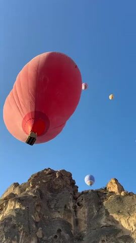 A low-angle shot of a large red hot air balloon dominating the foreground. It is partially inflated and tilted, with the basket visible at the bottom. In the background, several smaller hot air balloons are visible against a clear blue sky. The largest of these has a pink and white pattern, and another is yellow. Below the balloons, rugged, tan-colored rock formations are visible, suggesting a mountainous or canyon landscape. The lighting indicates it may be early morning or late afternoon. The overall scene is serene and scenic.