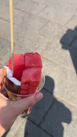 Here is a description of the video:

The video shows a person holding a clear plastic cup filled with fresh fruit, including watermelon, pineapple, kiwi, strawberries, and coconut. The camera then follows a woman walking down a pedestrian street. The woman has long, wavy brown hair and is wearing a white button-down shirt and black pants. She is carrying a white purse with a chain strap. The street is paved with light-colored stone and lined with buildings and shops. People are walking in the background. The sky is blue and sunny.