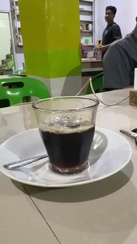 This media file shows a close-up of a glass of black coffee with foam on a white saucer. A spoon rests on the saucer. The coffee is placed on a table in what appears to be an indoor setting, possibly a cafe or eatery. In the background, a person is visible, wearing a dark t-shirt. Another person, partially visible, is seated to the right, wearing a gray t-shirt. A pillar painted with yellow and green stripes stands behind the coffee. Green chairs are also visible. The lighting suggests it could be daytime or evening. The scene is set in Manyang Cut, Indonesia. There is no visible text.