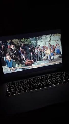 The video shows a laptop with a movie playing on its screen. The movie appears to be a historical drama or fantasy, with a large group of people dressed in ancient-looking clothing gathered outdoors in a rocky, natural setting. The scene on the laptop screen changes to focus on two men with beards, one of whom is speaking. The laptop is open, and its keyboard is illuminated. The overall lighting is dim, suggesting the video was recorded in a dark room.