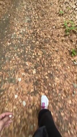 The video shows a first-person perspective of someone walking on a leaf-covered path through a forest. Two people are walking ahead, one with a blue backpack and trekking poles, and the other in a red top. The ground is covered with brown leaves, and there are green trees and foliage on either side of the path.