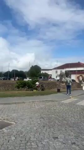 The video shows a man playing drums on the side of a street. He is wearing a white shirt and red shorts. He is sitting on a stool behind a drum set. There are cars and people in the background. The ground is paved with cobblestones. There is a short stone wall covered in greenery behind the drummer. The sky is blue with some clouds.