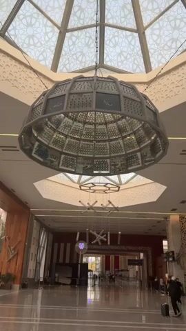 The video shows the interior of a large building, possibly an airport or a convention center, with a high, ornate ceiling. A large, domed chandelier hangs prominently in the center of the frame, adorned with geometric patterns and what appear to be inscriptions. Below it, a more modern, geometric light fixture is suspended. The floor is polished and reflective, and the walls are decorated with patterned wallpaper. In the distance, several people can be seen walking, some with luggage. The lighting is bright, suggesting daytime. The overall impression is one of grandeur and spaciousness.