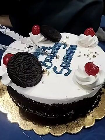 A birthday cake with white frosting and dark chocolate sides is decorated with blue icing that spells out "Happy Birthday Sharnu." The cake is adorned with dollops of white frosting, red cherries, and Oreo cookies. Gold sprinkles are scattered across the top. A knife is visible, partially inserted into the cake, indicating it is being cut. The cake rests on a gold scalloped cake board, which is placed on a dark blue surface. A person's hands and arms are partially visible in the upper portion of the frame, suggesting they are cutting the cake. The overall scene appears to be a celebration.