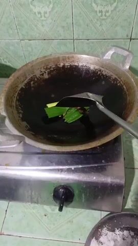 This media file shows a close-up, overhead view of a wok on a stove in an Indonesian kitchen. The wok contains a dark, oily substance and several green pieces of what appear to be pandan leaves, being stirred by a metal spatula. The background features a green tiled wall with a repeating geometric pattern. The lighting suggests it is daytime, and the setting is domestic. The overall impression is a preparation of food or an aromatic infusion. There is no visible text in the media file.