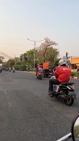 In a medium shot, the camera captures a scene on a paved road in Klurak, Indonesia. The time of day appears to be late afternoon or early evening, as indicated by the warm, soft lighting and the hazy sky. The weather seems pleasant, with clear skies and gentle breezes that rustle the leaves of the trees lining the road.

The primary subjects are several individuals riding motorcycles. In the foreground, a person in a red shirt with white lettering and a white helmet rides a scooter. Following closely behind them is another scooter carrying two people, one wearing a striped shirt and the other a dark helmet. Further down the road, another motorcycle rider is visible, also wearing a red shirt.

Other objects in the scene include a black flatbed truck carrying a large, orange, tarp-covered object. The truck is positioned in the middle of the road, moving in the same direction as the motorcycles. The road itself is asphalt, with a white dashed line marking the lanes. The sides of the road are bordered by a sidewalk and then lush greenery, including palm trees and other tropical foliage. In the distance, some buildings are partially visible.

The activity taking place is simply traveling on the road, likely commuting or delivering goods. The overall mood is peaceful and ordinary, depicting everyday life in a tropical urban setting. The visible text on the red shirt appears to be a logo or brand name, but it's too blurry to read clearly. The license plate on the scooter in the foreground is also unreadable.