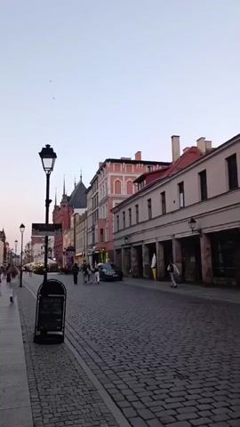 The video shows a cobblestone street in what appears to be a European town or city. The street is lined with old buildings, some of which have arched walkways on the ground floor. There are several people walking on the sidewalk and in the street. A street lamp is visible on the left side of the screen, along with a signpost. In the distance, there are more buildings and some trees. The sky is clear and the light suggests it is either early morning or late afternoon. The overall atmosphere is calm and peaceful.