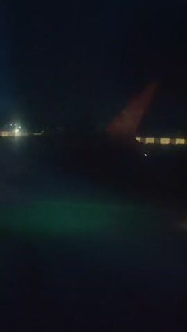 The video is a shaky, blurry, nighttime view from an airplane window. The focus is on the wingtip of the plane, which is orange with a white logo. The wingtip is silhouetted against a dark sky. In the background, there are lights on the ground, suggesting an airport tarmac. The lights are out of focus and appear as streaks of light. The video appears to be of the plane taxiing on the runway or preparing for takeoff.